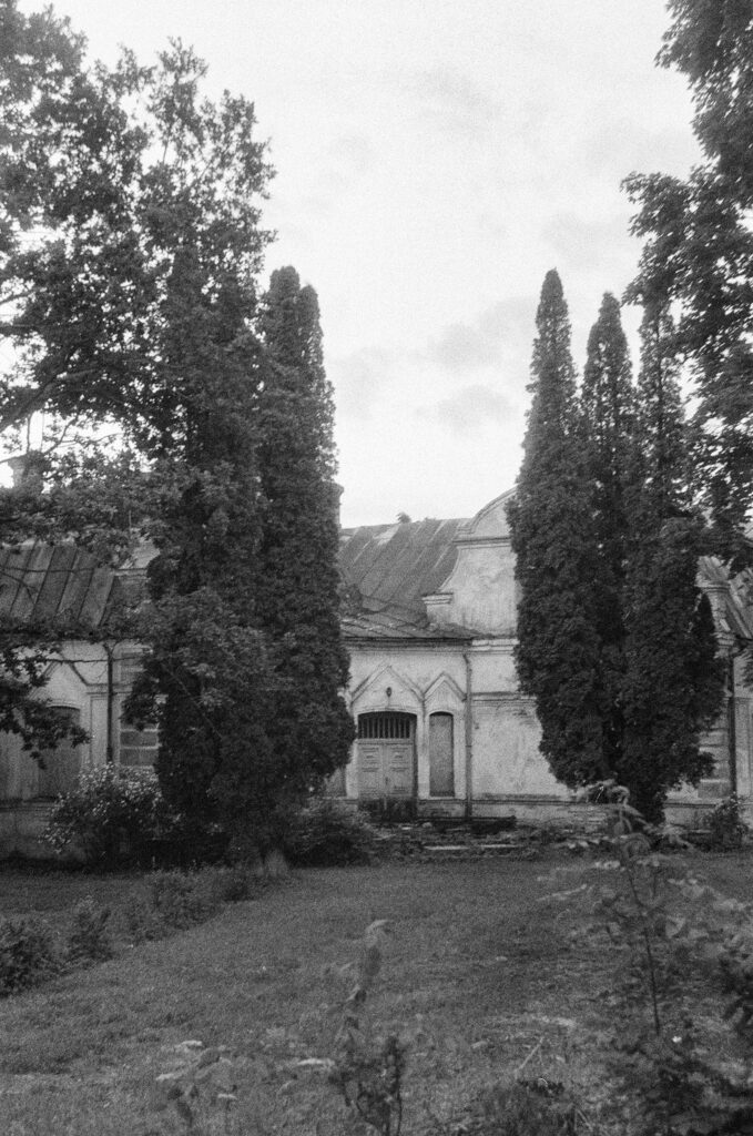 Old manor house framed by tall conifer trees in a grainy black-and-white photograph.