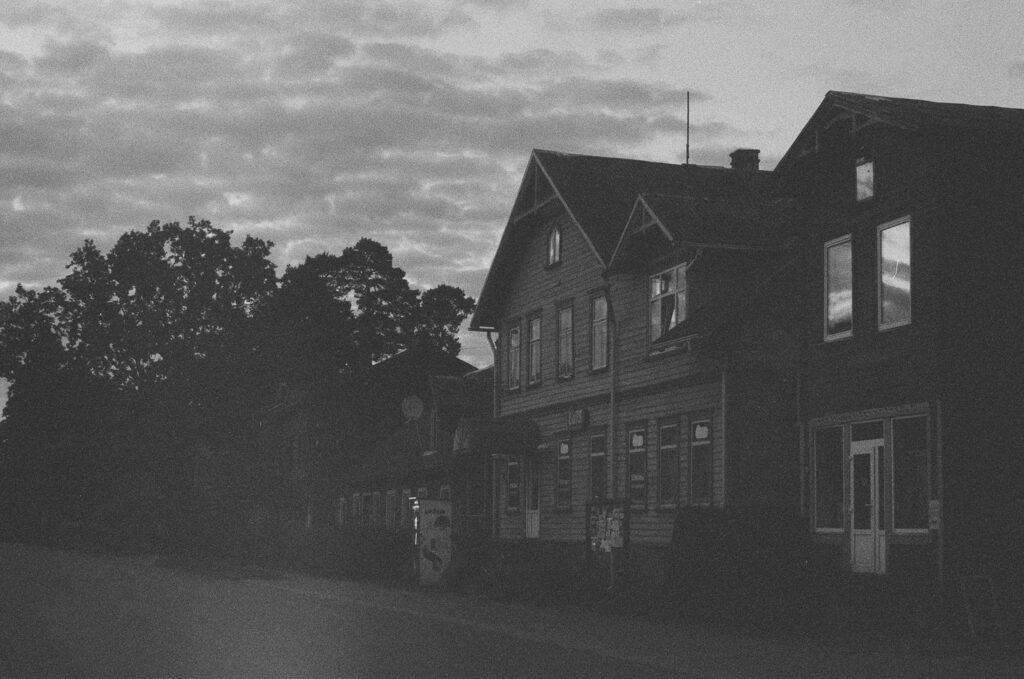 Dark wooden houses in Ainaži at dusk, with a cloudy sky reflected in the windows.