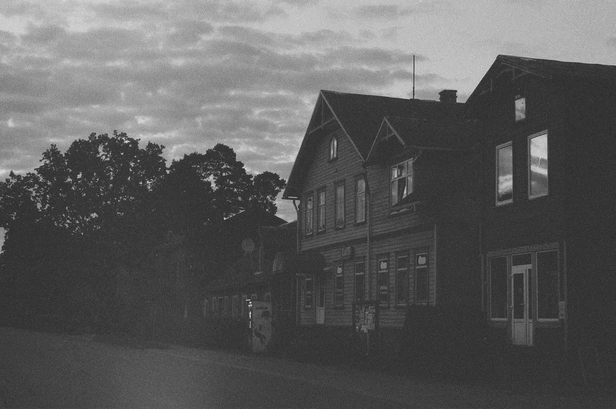 Dark wooden houses in Ainaži at dusk, with a cloudy sky reflected in the windows.