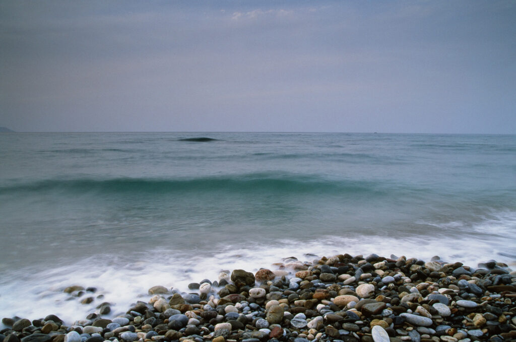 Long-exposure waves washing over a pebble beach beneath a quiet blue-grey Aegean sky.