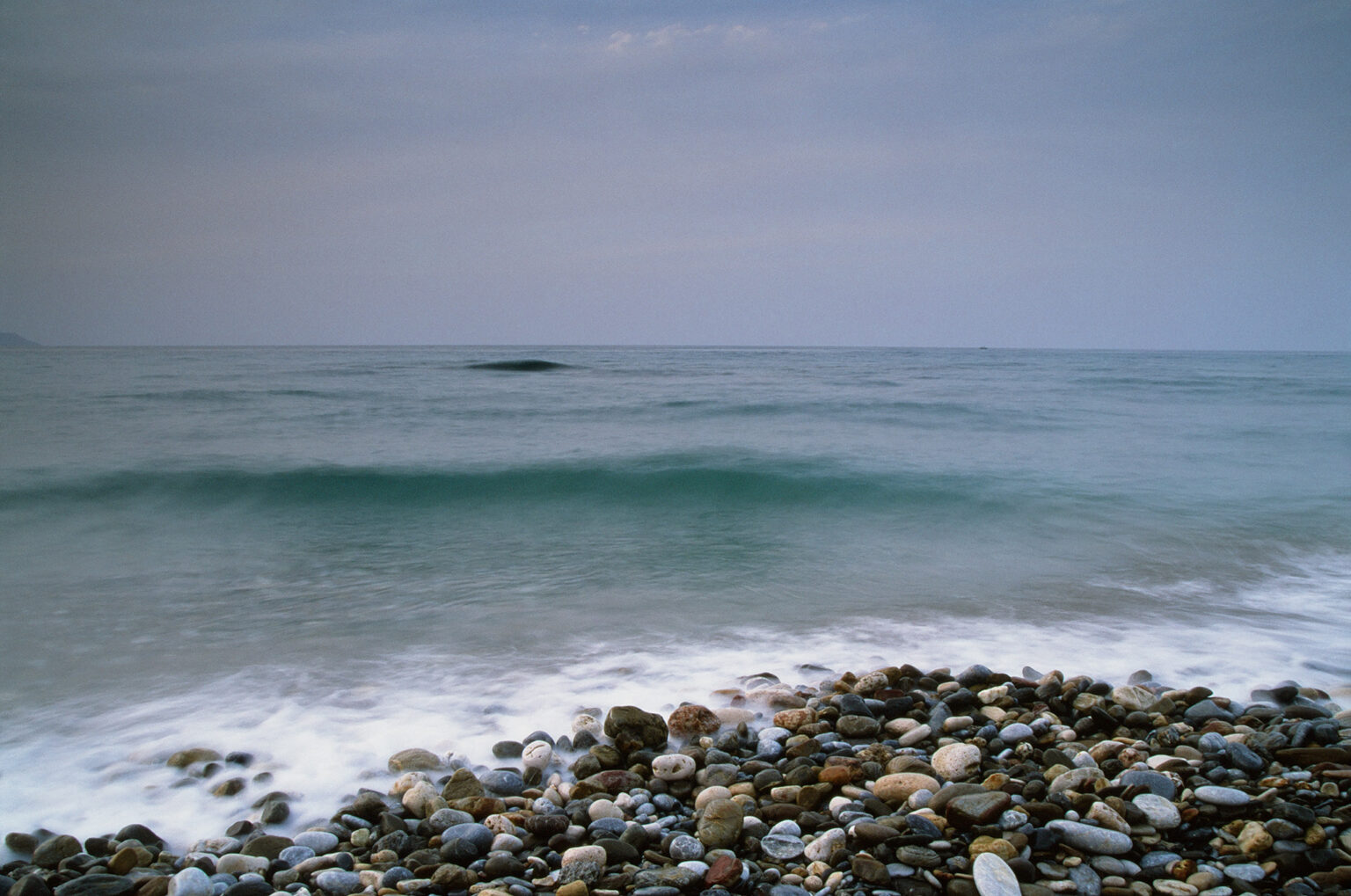 Long-exposure waves washing over a pebble beach beneath a quiet blue-grey Aegean sky.